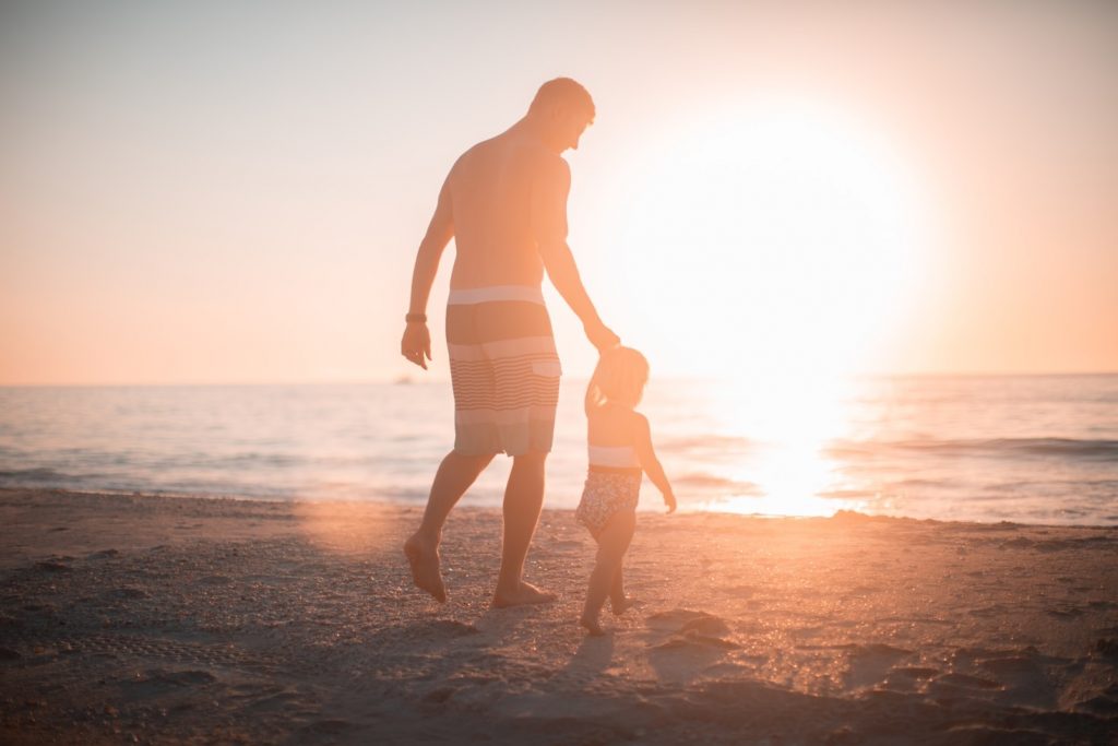 Ein Mann geht mit einem Kind am Strand entlang, die Sonne steht tief und strahlt warmes Licht – Symbol für Nähe und bewusste Momente im Vatersein.